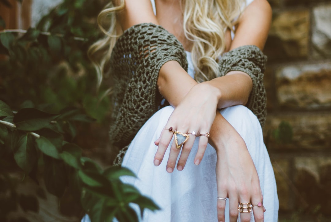 A person with long blonde hair, wearing a white dress and knitted shawl, sits outdoors with their arms crossed, showcasing stylish jewelry and several rings—perfect inspiration for jewelry gifts for him.