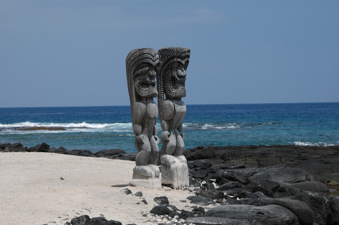 Two carved wooden statues stand on a sandy area near black rocks, overlooking the ocean under a clear blue sky—a perfect scene for USA travel and those seeking the ultimate travel experience.