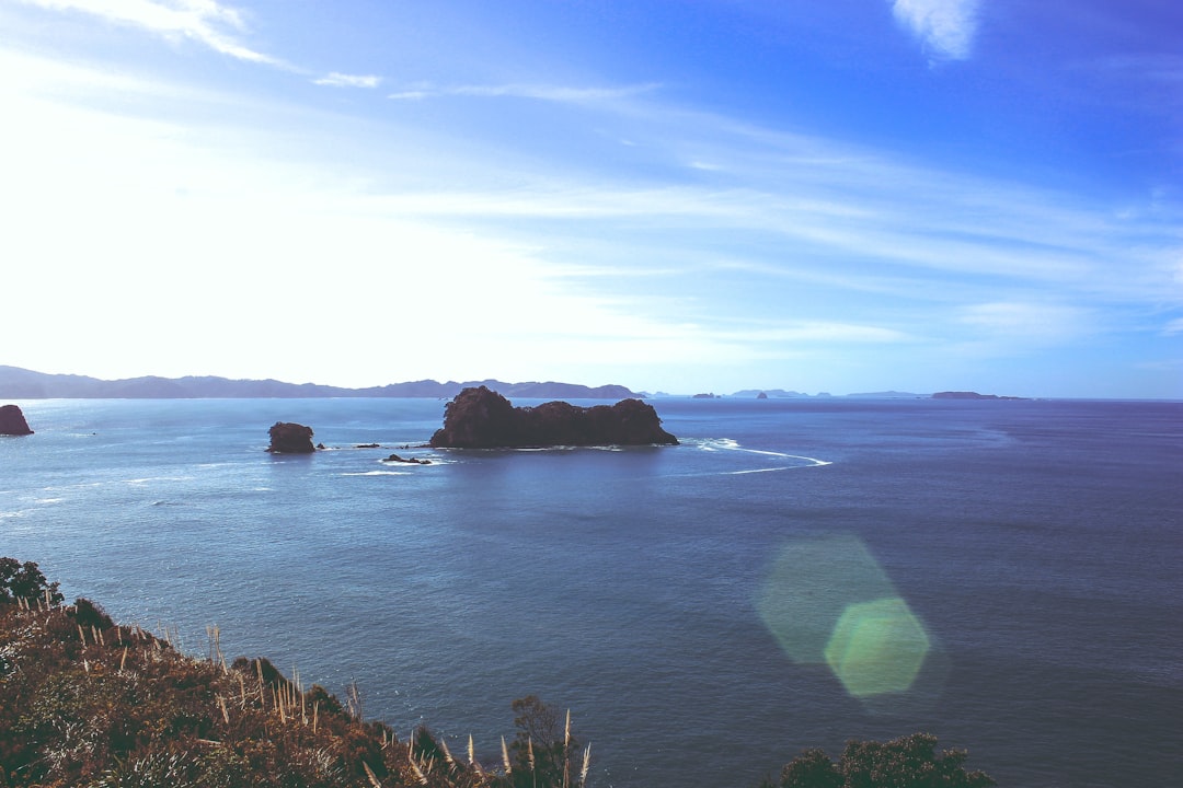 View of a small rocky island surrounded by blue ocean under a clear sky, with distant land on the horizon—a scene perfect for New Zealand adventures or capturing memories from your tailored trip. Lens flare adds to the magic in the foreground.