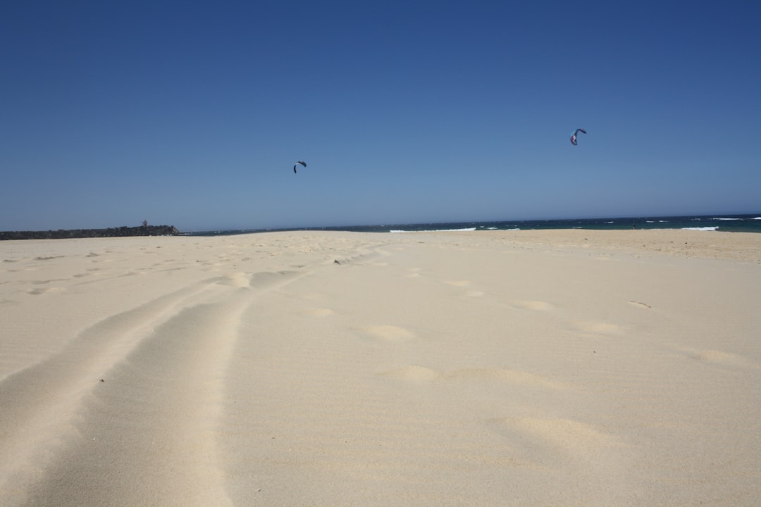 Wide sandy beach under a clear blue sky, with two distant kite surfers above the horizon. Tire tracks curve across the sand in the foreground—perfect for those seeking the Best Down Under Experience on a luxury travel Australia adventure.