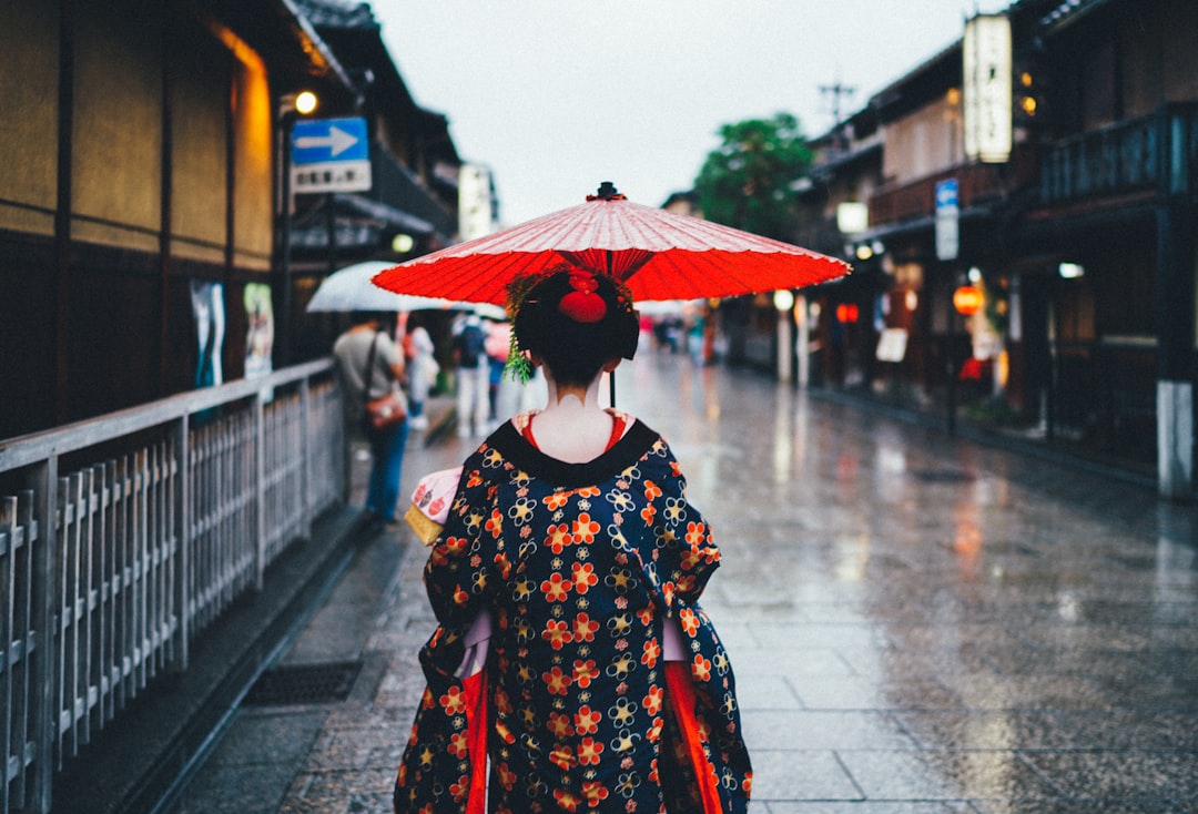 A person in traditional Japanese clothing holds a red umbrella while walking down a wet street lined with old-style buildings, capturing the timeless beauty of the Land of the Rising Sun.