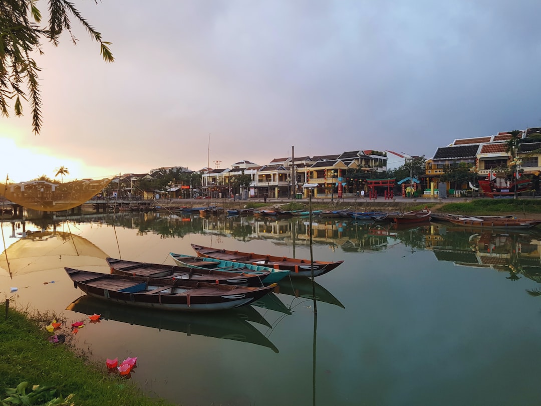 Several wooden boats are docked on a calm river, where buildings and houses line the opposite bank under a cloudy sunset—a scene reminiscent of historic European river cruises.