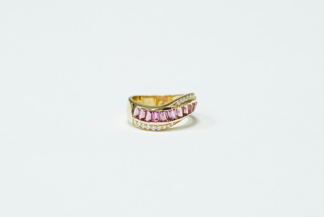 Gold ring featuring a row of pink rectangular luxury gemstones bordered by two rows of small white stones, making it a standout among coveted jewels, displayed on a plain white background.
