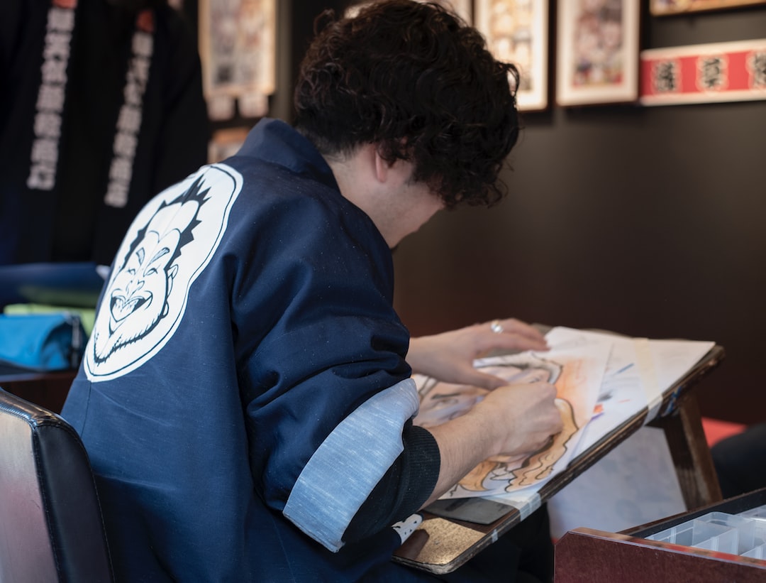 A person with curly hair sketches on a large pad while sitting at a desk in a blue jacket with a cartoon face on the back. Framed art pieces inspired by Japanese craftsmanship hang on the wall in the background.
