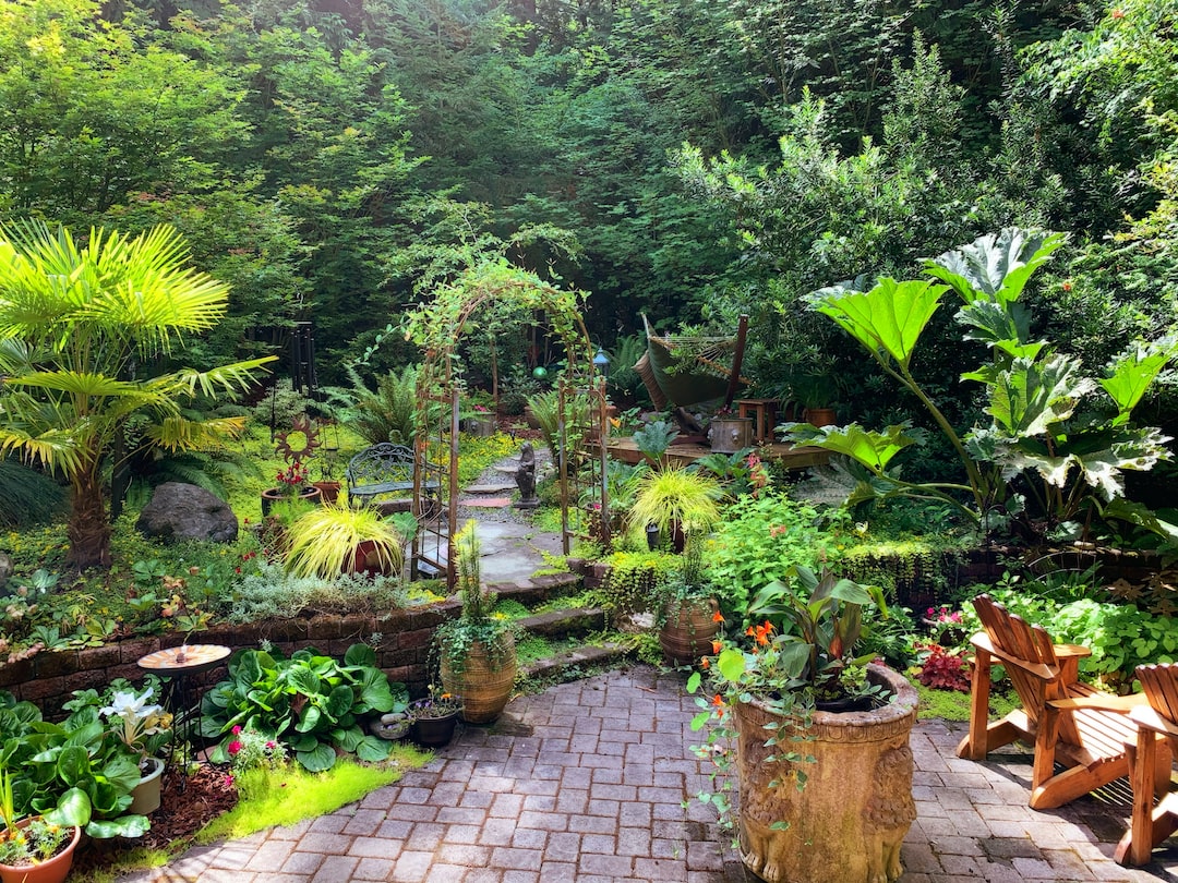 A lush, green garden with various plants, potted flowers, a stone path, wooden chairs, and a hammock reflects thoughtful landscape design, surrounded by dense trees in the background.