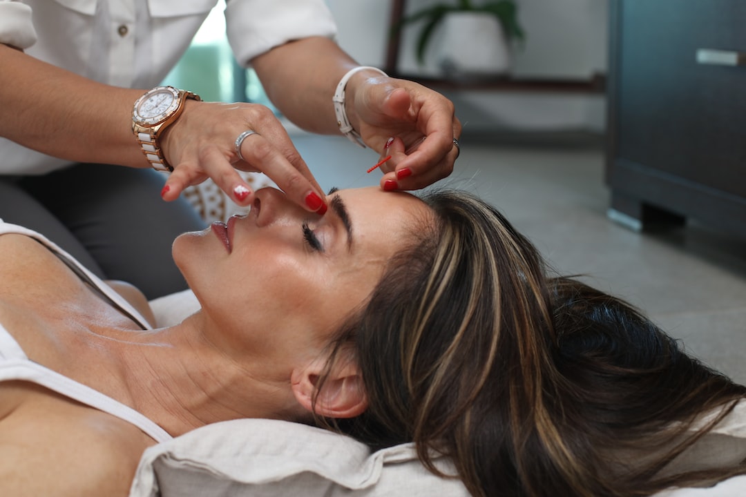 A woman lies on a cushion with her eyes closed while another person shapes her eyebrows using tweezers in a serene setting inspired by luxury herbal medicine.