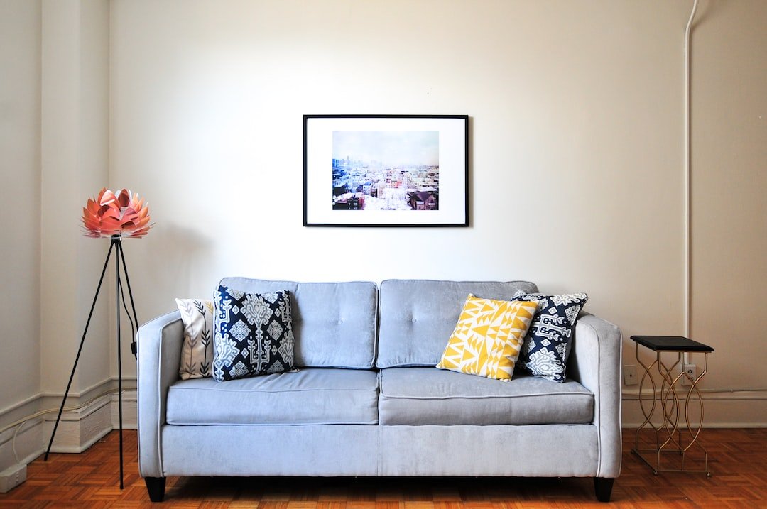 A light gray sofa with patterned and yellow pillows sits against a plain wall, beneath a framed cityscape photo reminiscent of an exclusive gallery. A pink floor lamp and a small side table are nearby.