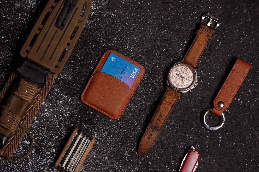 A brown luxury leather watch, cardholder with a blue Visa card, keychain, knife, and multitool showcase fine leather craftsmanship on a dark, textured surface.