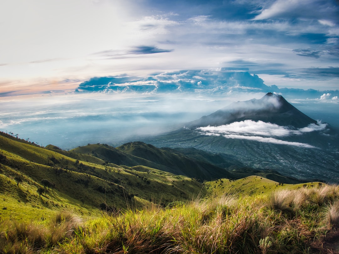 A grassy hillside overlooks a distant, cloud-covered mountain under a partly cloudy sky, evoking the marvel of a Horological Wonder from the Ulysse Nardin Freak Collection.