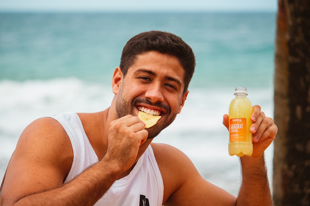 A man in a white tank top smiles while holding a potato chip to his mouth and a bottle of yellow juice by the ocean, enjoying the day after applying the best face sunscreens to protect skin from the sun.