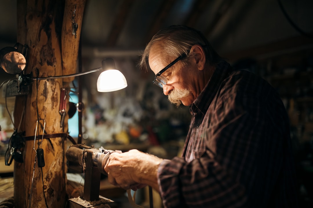 An older man wearing glasses works intently with tools at a wooden workbench in a dimly lit workshop, reminiscent of traditional craft workshops.