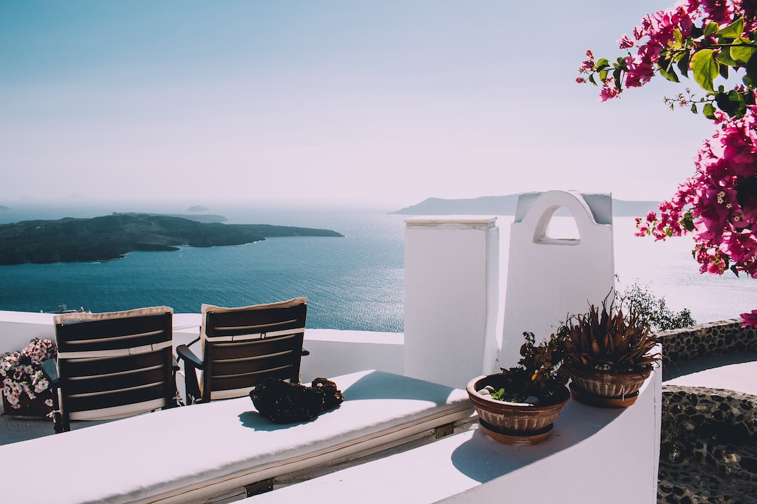 Two lounge chairs and potted plants on a white terrace overlook a calm blue sea, with distant islands and pink flowers—an inviting scene perfect for high-end health escapes or tranquil wellness retreats.
