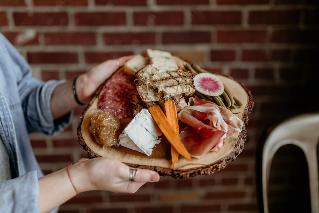 Person holding a wooden platter with assorted charcuterie items, featuring artisan cheese, cured meats, bread, pickles, carrots, and sliced radish, against a brick wall background.