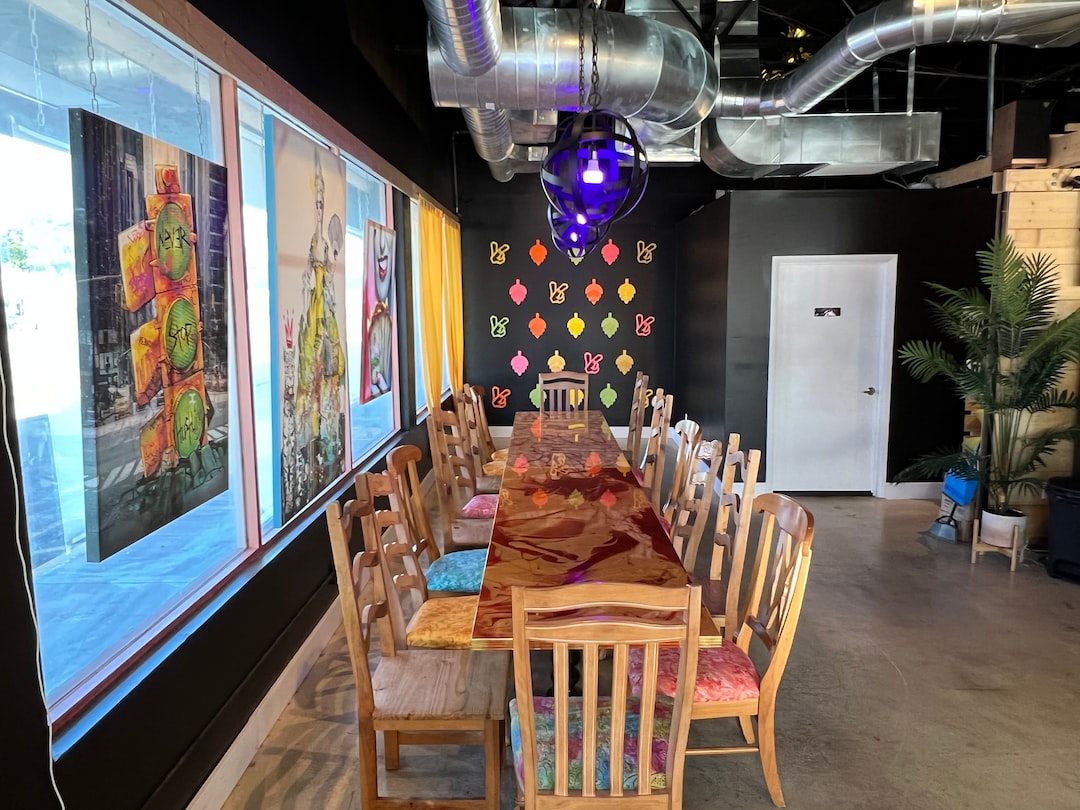 A long, custom-made wooden dining table with mismatched chairs is set in a colorful, modern room adorned with artwork and exposed ductwork on the ceiling, highlighting unique craftsmanship.