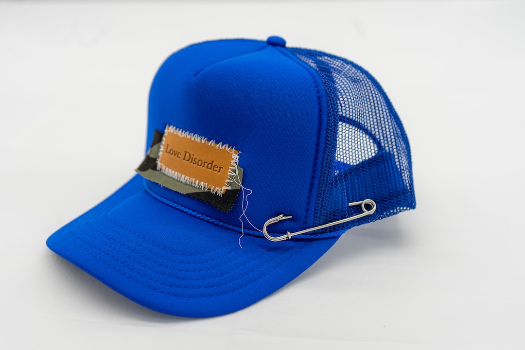 A blue mesh trucker hat inspired by avant-garde fashion, featuring a "Love Disorder" patch and a large safety pin for an edgy twist.