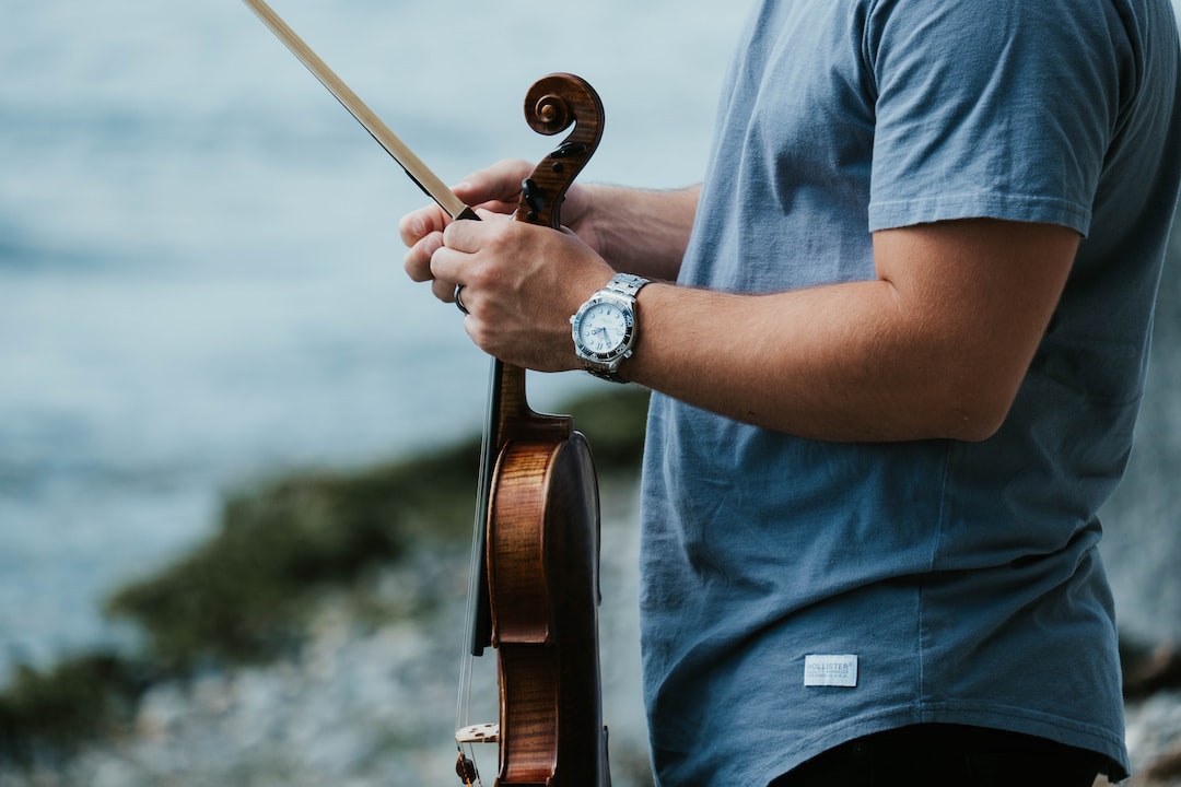 A person in a blue shirt holds a violin and bow outdoors, embodying the design philosophy of precise artistry found in Bell & Ross instrument watches, with a blurred background of water and shoreline.