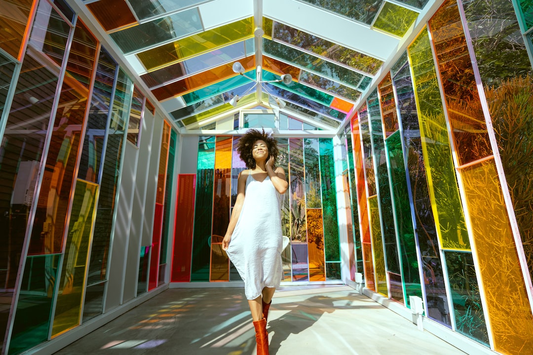 A person in a white dress and red boots stands in a vibrant, colorful glass-paneled structure, with light streaming through the eclectic mix of multicolored panels.