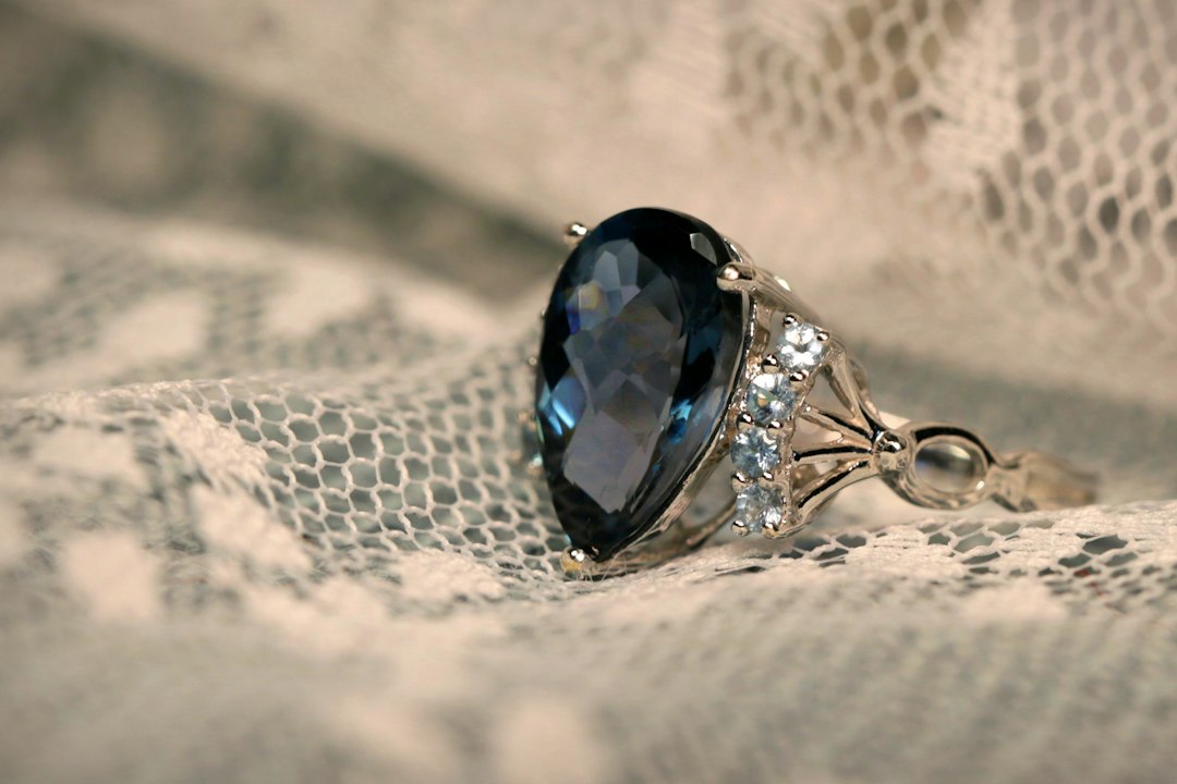 A silver ring with a large pear-shaped dark blue gemstone and smaller clear stones rests on white lace fabric, capturing the timeless beauty of gemstones.