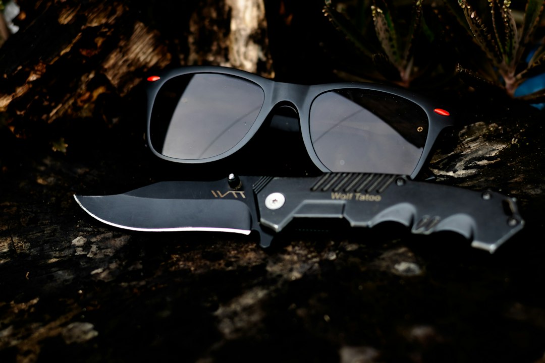 A pair of black sunglasses showcasing the latest designer eyewear trends and a black folding knife labeled "Wolf Tattoo" rest on a dark, textured surface.