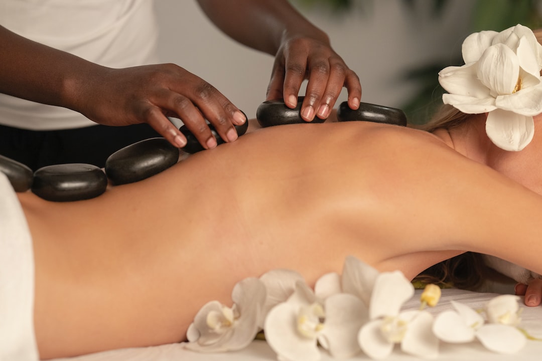 A person enjoys a soothing hot stone massage on their back in a tranquil spa setting, surrounded by white orchids, with one delicate flower tucked in their hair.