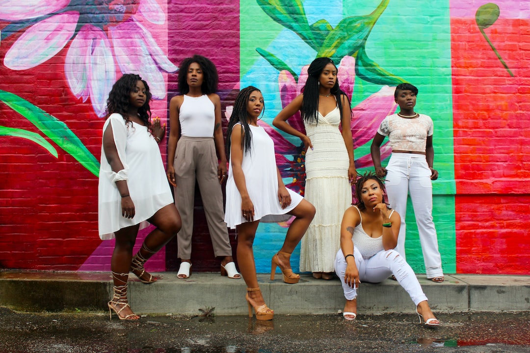 Six women pose confidently in front of a colorful mural with large floral designs. Wearing coordinated white outfits and subtle wearable tech, they exude a sense of future luxury as they stand on the wet pavement.