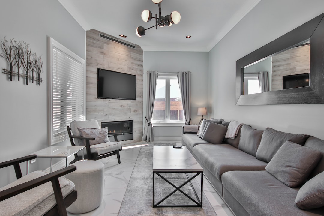 Modern living room with gray sofa, large wall mirror, mounted TV above fireplace, armchairs, coffee table, and window with curtains letting in natural light—perfect for luxury living in the heart of New York City.