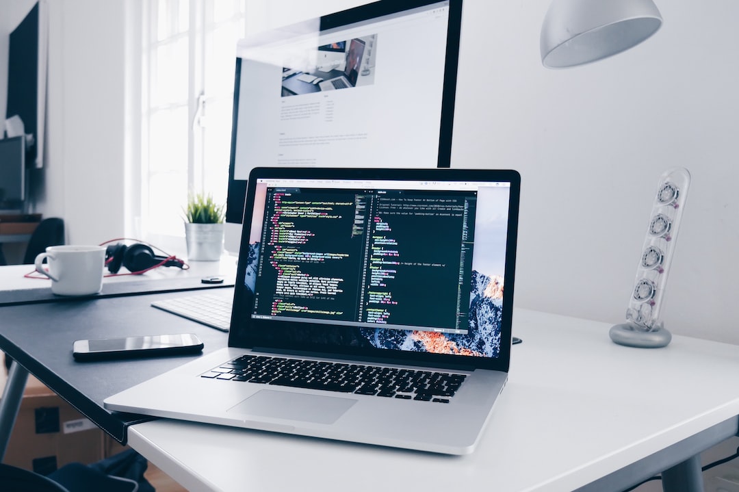 A laptop displaying code sits on a white desk with a large monitor, headphones, and other office items in a modern workspace—a testament to the impact of technology on today’s work environments.
