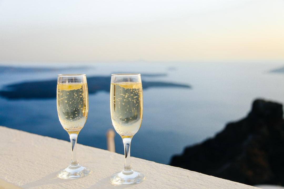 Two champagne flutes filled with sparkling wine sit on a white ledge overlooking a calm sea and distant cliffs at sunset—a scene reminiscent of moments found at luxury wellness retreats.