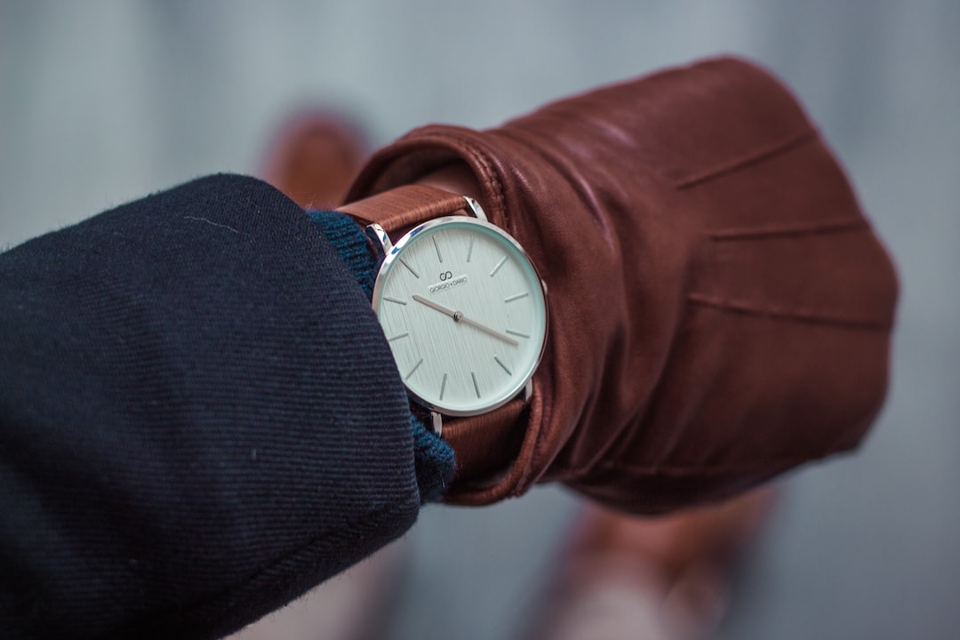 A person wearing a brown leather glove and a dark coat looks at a silver wristwatch with a white face and minimalistic design, embodying the art of mastering time.