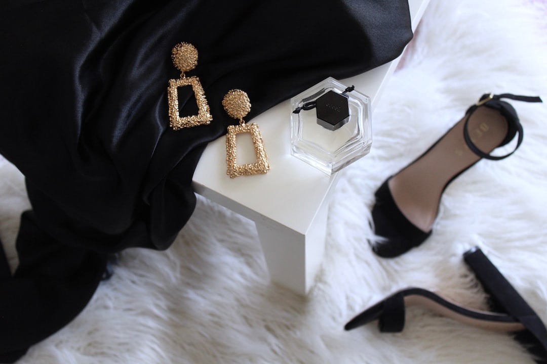 Gold geometric earrings crafted from precious metals, a bottle of perfume, and black high heel shoes are arranged on a white table with black fabric, set atop a plush white furry surface.