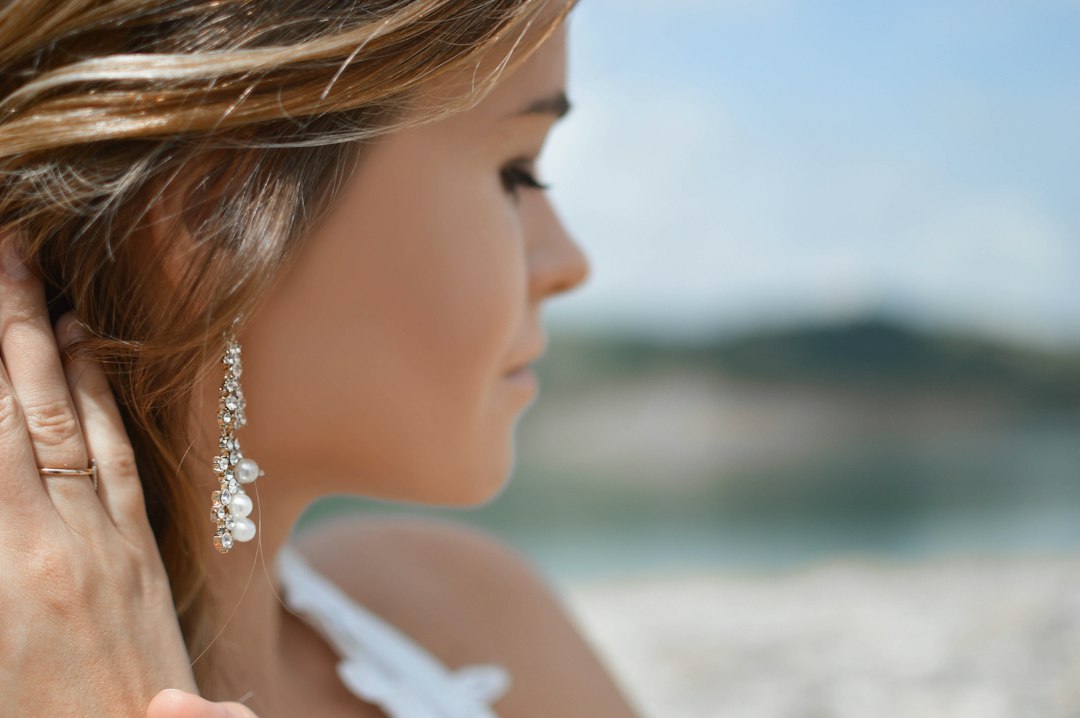 Close-up of a woman with long brown hair wearing a pearl and crystal earring, lightly touching her ear—a chic touch of zodiac jewelry—set against a blurred outdoor background.