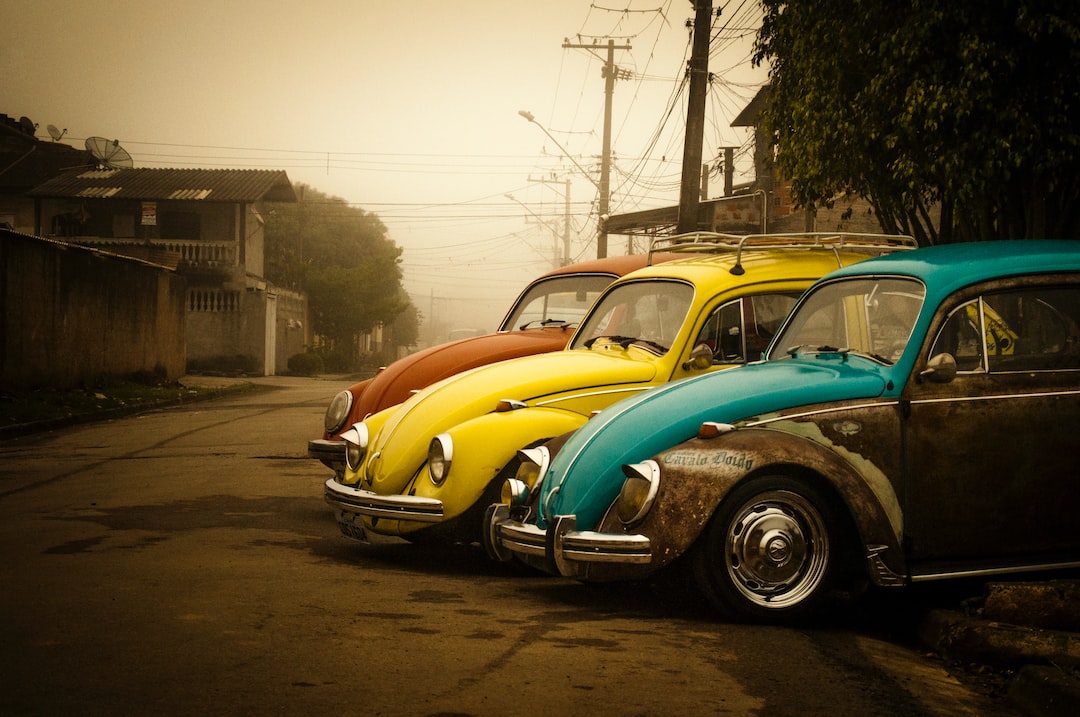 Four iconic vintage Volkswagen Beetles in blue, yellow, orange, and brown are parked in a row on a quiet, foggy street beside houses and power lines.