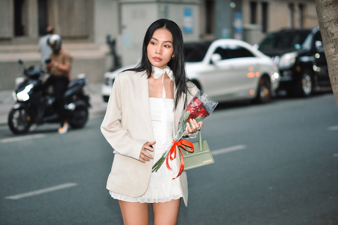 Woman in a white outfit and beige blazer, reminiscent of Ralph & Russo luxury evening wear, holds a bouquet of red roses on a city street with cars and a person on a scooter in the background.