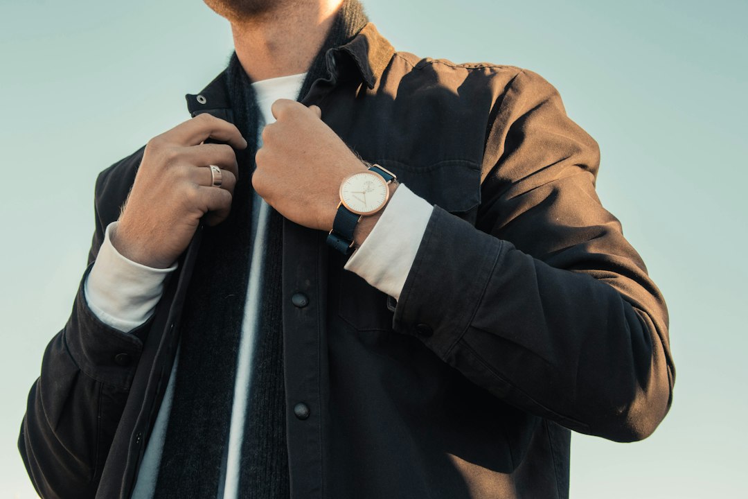 A person adjusts their dark jacket with elegance, sporting a Swiss watch and a ring on their left hand, set against a light sky in the background.
