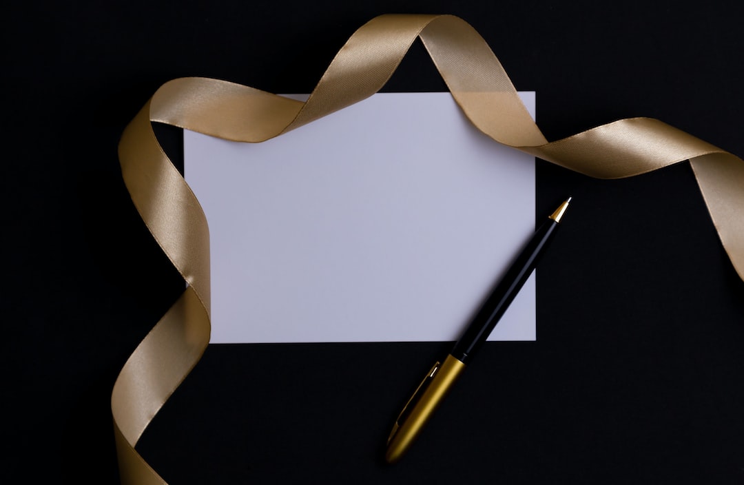 A blank white card with a gold pen, perfect for luxury pen collecting enthusiasts, rests on a black surface, partially bordered by a gold satin ribbon.