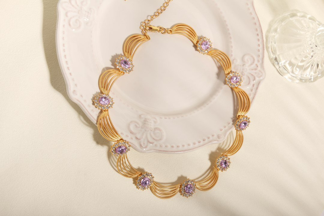 A gold necklace with purple gemstone accents is displayed on an ornate white plate beside a clear glass object, offering a glimpse of refined style and elegance, all set on a light-colored surface.