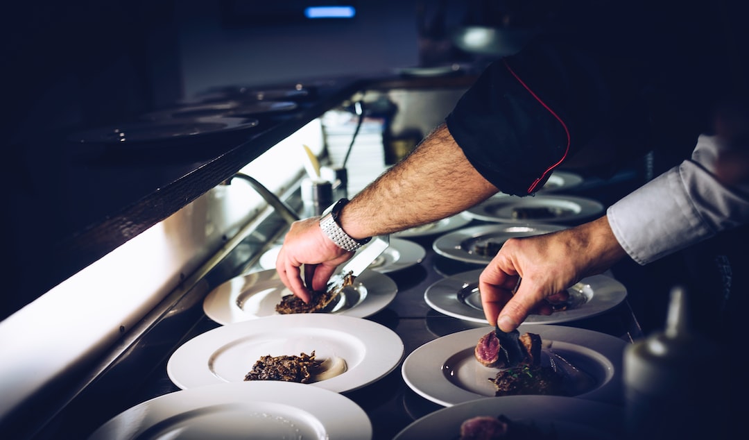 A chef’s hands, inspired by celebrity cuisine, artfully plate gourmet food on several white dishes in a restaurant kitchen.