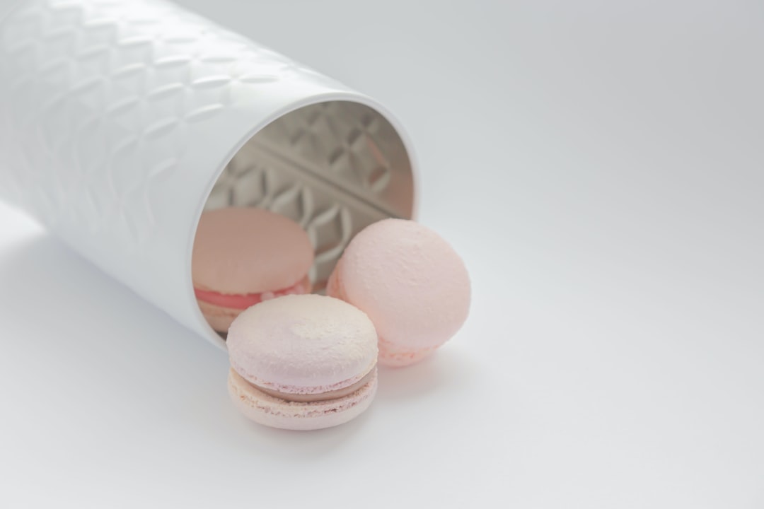 Three pastel-colored macarons, crafted with gourmet baking expertise, spill out of a white textured container onto a light surface.