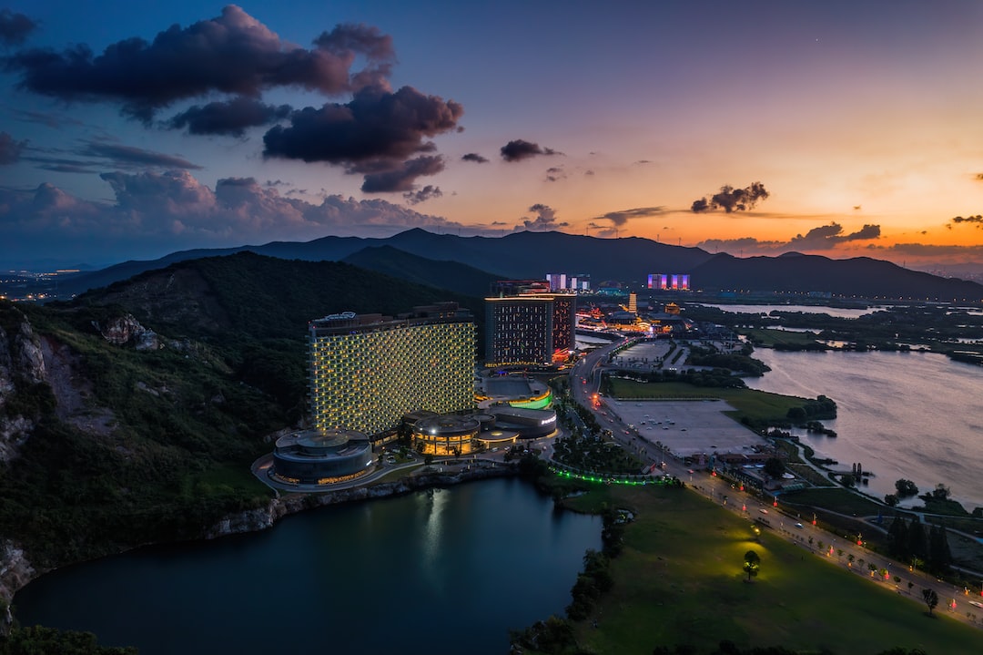 Aerial view captured by high-end drones shows a brightly lit hotel complex by a lake at sunset, surrounded by mountains and a distant cityscape beneath a colorful sky—ideal for personal use in travel memories.