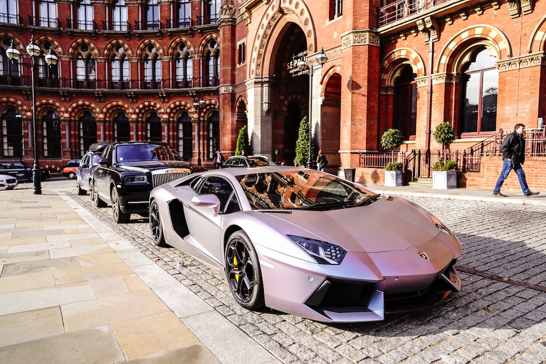 A matte silver Lamborghini, the epitome of a luxury car, is parked on a cobblestone street in Moscow, in front of an ornate red brick building, with other high-end cars lined up behind it.