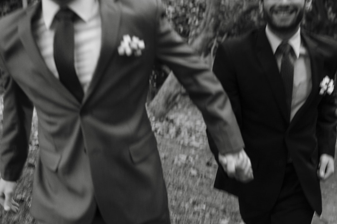 Two men in suits, showcasing classic tailoring, hold hands and walk outdoors, partially out of frame with blurred motion and trees in the background.