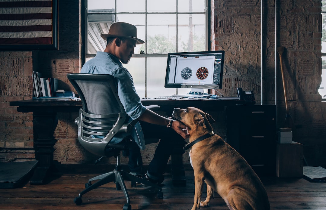 A man in a hat sits at a desk with a computer, petting a brown dog in a rustic office with a large window and framed flag on the wall, his wrist adorned with one of the latest designer watches, adding timeless elegance to the scene.