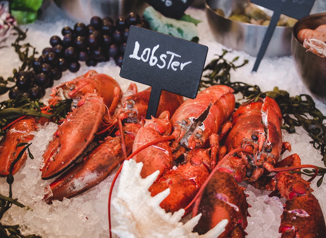 Two whole lobsters are displayed on ice at a seafood market, with a sign labeled "Lobster" above them. Grapes and other exclusive seafood are visible in the background, highlighting a selection perfect for gourmet seafood dishes.