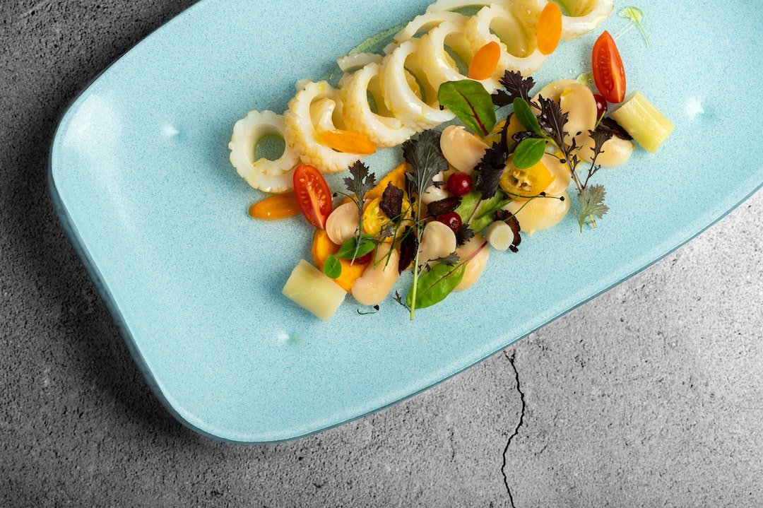 A must-try for any connoisseur, this light blue plate features arranged calamari rings, cherry tomatoes, leafy greens, white beans, cheese cubes, and small herb garnishes on a gray surface.