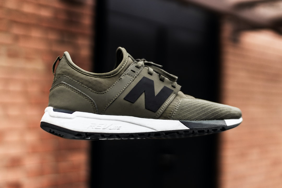 A green and black athletic sneaker with white sole featuring a large "N" logo, designed by bespoke shoe designers, photographed in mid-air against a blurred brick wall background.