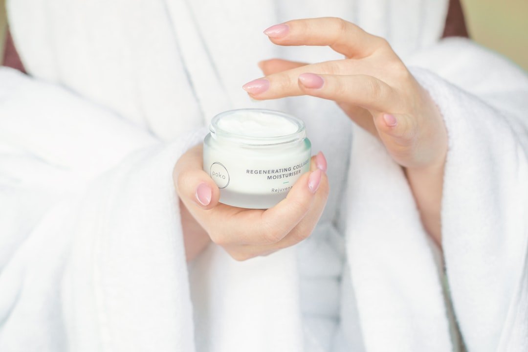 A person in a white robe holds a jar of the best anti-aging collagen moisturizer and is about to apply the cream with their fingers, showcasing premium skincare products.