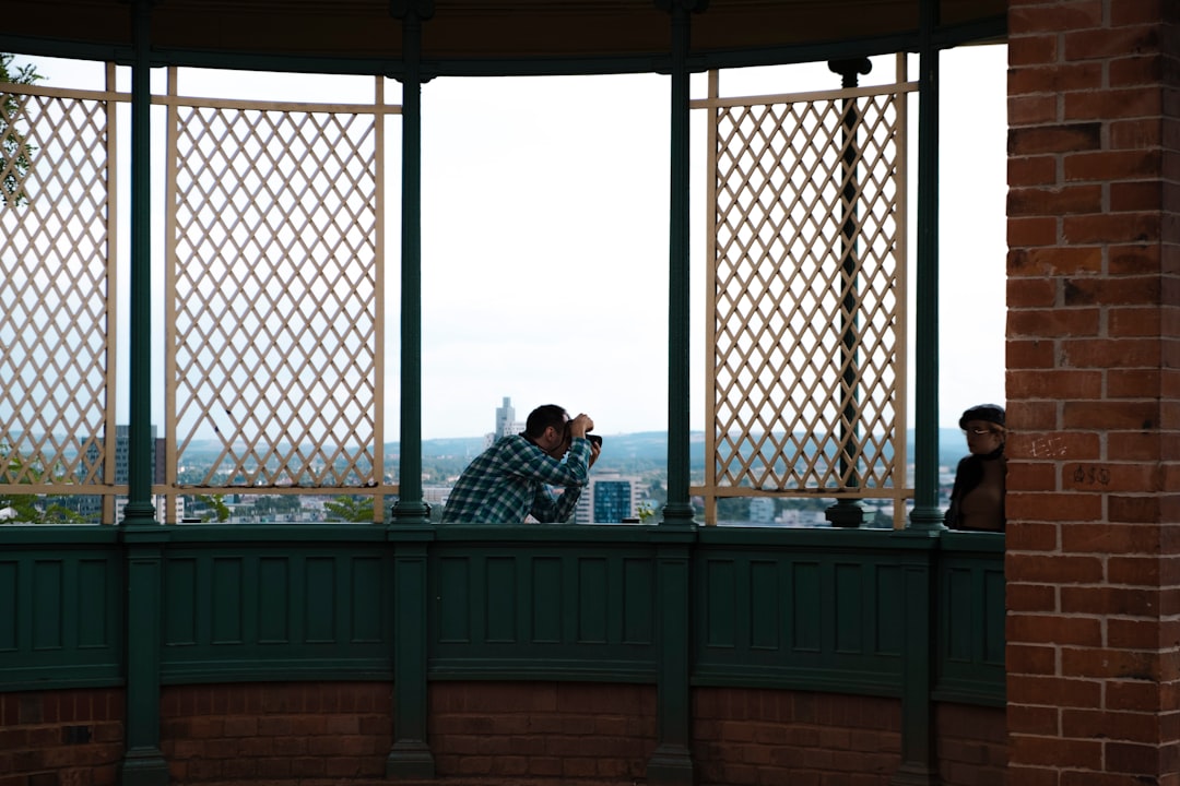 A person in a checkered shirt takes a photo of another on a balcony with lattice panels, overlooking a cityscape—capturing the moment just like Beauty Apps enhance your look for stunning memories.