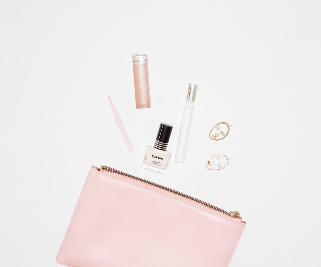 A pink zipper pouch with top picks in beauty products—nail polish, lip gloss, perfume bottle, pink tweezer, and two gold hoop earrings—arranged on a white background. All items are curated for under 20 Euros.