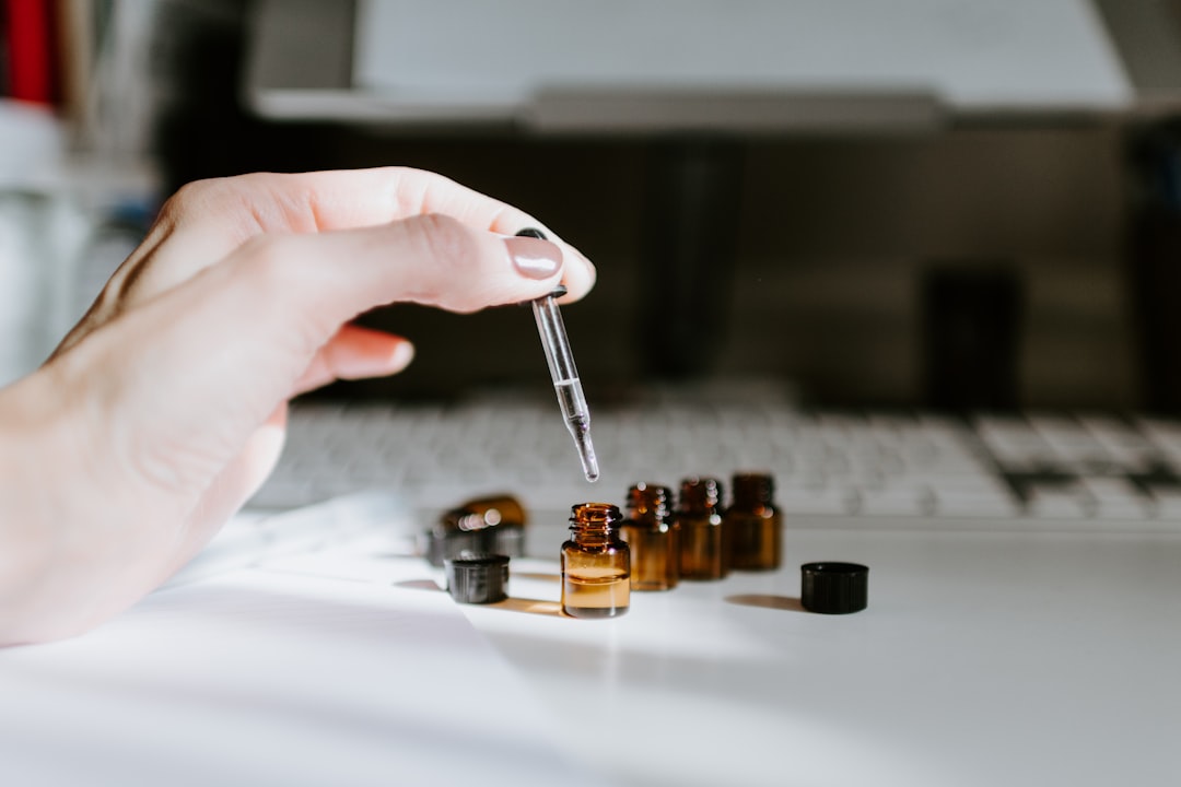A hand holds a glass dropper above a small brown glass bottle of facial oils, with several similar bottles and black caps on a white desk—perfect for achieving glowing skin.