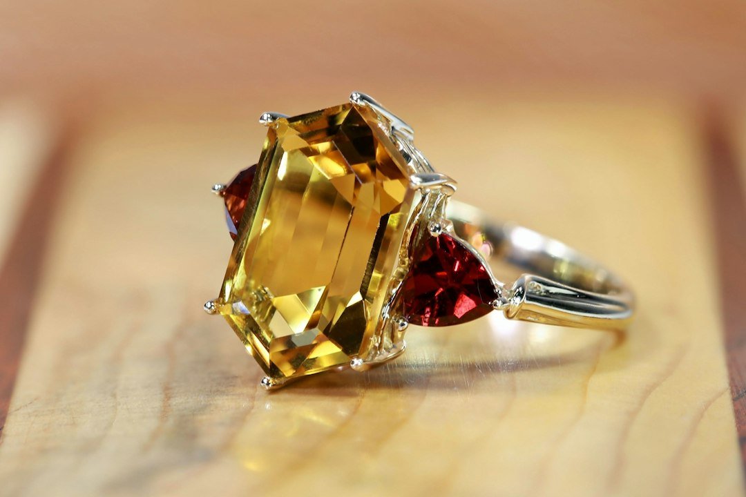 A gold ring with a large rectangular yellow gemstone flanked by two smaller red gemstones—nature’s treasures in exquisite gemstones—displayed on a wooden surface.